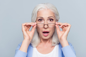 Close up portrait of stylish, aged, charming, surprised, shocked woman holding eyelets peek out glasses with wide open eyes and mouth over grey background