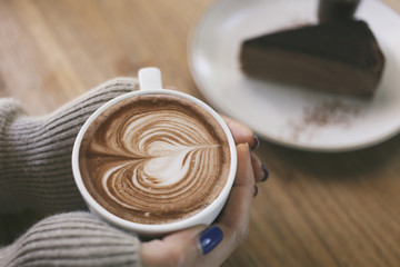 Top view female hands holding a cup of hot coffee with heart shaped latte art and plate of chocolate cake on the table.