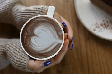 Top view female hands holding a cup of hot coffee with heart shaped latte art and plate of chocolate cake on the table.