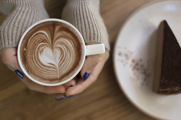 Top view female hands holding a cup of hot coffee with heart shaped latte art and plate of chocolate cake on the table.