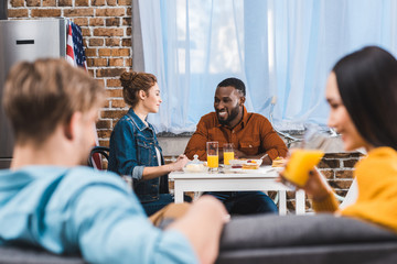 Fototapeta premium young smiling multiethnic couple sitting at table and friends drinking juice on foreground
