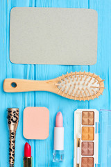 Makeup items on blue wooden background. Decorative cosmetics and natural hair brush, top view. Empty paper card and cosmetics products.