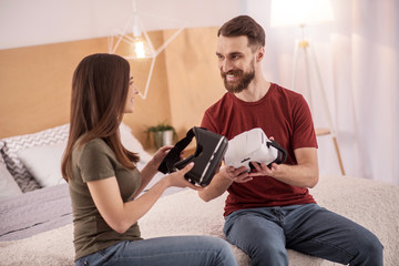 New tech. Attractive positive energetic couple sitting on the bad while looking at each other and holding VR glasses