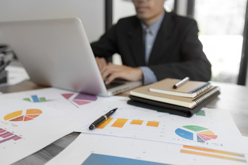 Close up view of notepads and pens on a work desk , in background hand of a man working on laptop computer.