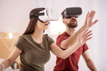 Virtual object. Pleasant glad charming couple stretching hands while putting on VR glasses and posing on the blurred background