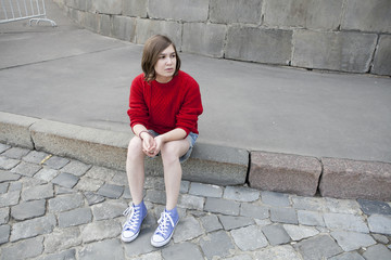 young girl in a red wool sweater and jeans shorts is sitting on the steps