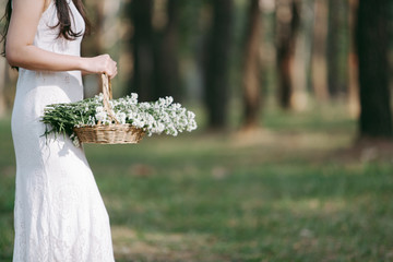 Female in white dress holding a basket with white flowers , side view in pine forest. with copy space.