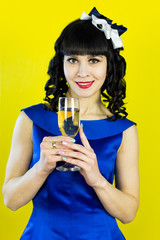 A girl with a glass of champagne smiles in front of the camera.. Beautiful brunette girl in a blue dress drinking champagne on a yellow background