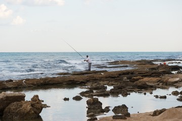 A man standing on sea rocks, fishing. Sea waves crashing on the rocks. Rocks reflected in the still water trapped between them.