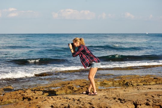 A young blonde woman wearing short shorts and plaid shirt taking pictures on a beach. Standing on sea rocks; sea, sky, clouds in background.