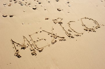 Mexico, with a hear shape for a dot over i, written in the sand. Some seashells and pebble laying around.