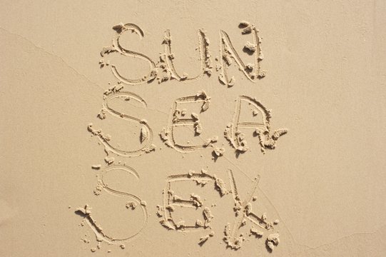 Sun, sea, sex written in the sand in all caps in a column.