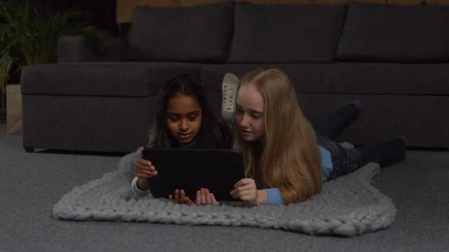 Adorable multi ethnic preteen girls browsing social networks on digital tablet while lying on the floor in domestic interior. Cute kids playing with touchpad and chatting while relaxing at home