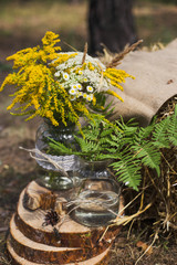 Beautifully decorated place in old autumn wood for wedding ceremony. Rustic style of elements of decor. Bright yellow flowers in unusual vases made of glass jars. Vertical color photography.