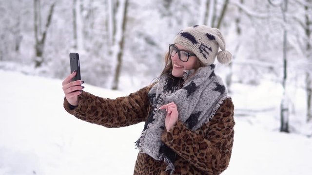 Attractive girl wears glasses have videochat by smartphone in winter snowy park