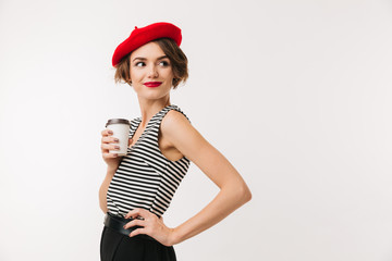 Attractive woman having french style wearing red beret posing on camera with takeaway coffee in paper cup and looking aside, isolated over white wall