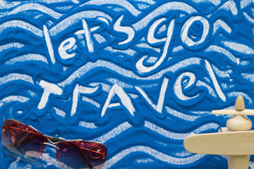 Let's Go Travel inscription on a blue colored sand with waves and travelling accessories, top view flat lay