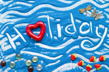 Holidays inscription on a blue colored sand with waves and decorative red plastic heart, top view, flat lay