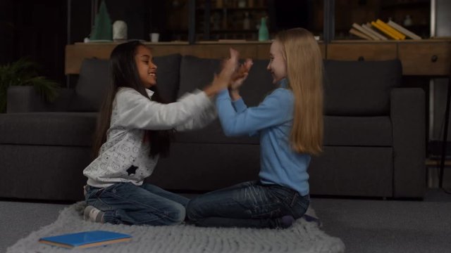Excited multiracial preteen girls playing clapping game while sitting on the floor at home. Side view. Smiling little friends having fun together while playing pat-a-cake game in cozy modern interior