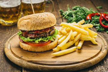 Delicious burger with beef cutlet, tomato, salad, parsley, sauce, pepper, onions, as well as French fries and beer. On a round wooden board