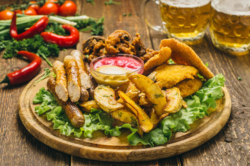 Potatoes, mushrooms in batter, chicken in batter, sausages on a round wooden board with lettuce, mustard, ketchup, parsley, pepper, tomatoes and beer