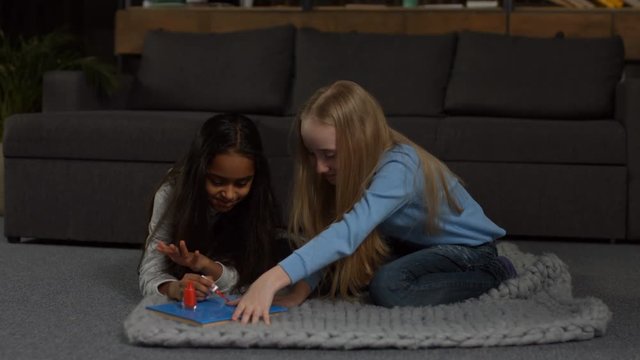 Cheerful mixed race little girl applying nail polish to her girlfriend while sitting on floor in domestic interior. Two adorable multi ethnic kids doing makeup and playing with cosmetic accessories
