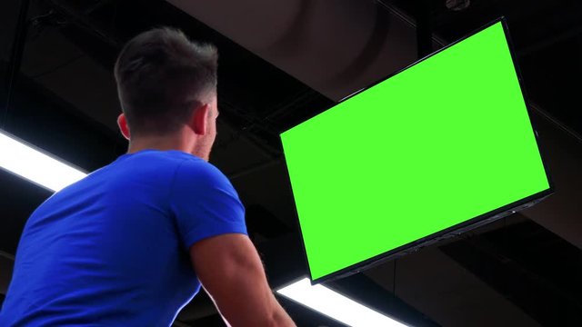 A young fit man trains on an exercise bike in a gym and watches a green screen - view from below