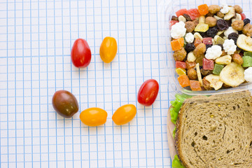 Naklejka na ściany i meble Fresh rye bread sandwich, candied fruit and nuts in a plastic box and a smiley face made of cherry tomatoes, top view, white squared paper background