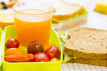 A green plastic lunch box with cherry tomatoes, carrot, a glass of juice and sandwiches close up, white squared paper background with copy space