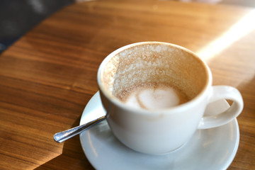 A white cup of hot coffee topping with milk foam leaf late art shape next by a small green plant on wooden table with morning lighting from window.