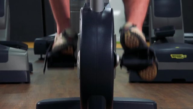 A fit man trains on an exercise bike in a gym - closeup on feet from behind