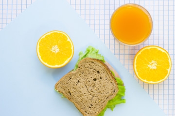 A rye bread sandwich, fresh orange and a glass of juice, top view, close up, blue and white squared paper background