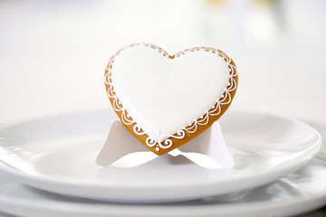 White plate,served with heart-shaped gingerbread cookie, covered with white glaze and decorated with pattern. Looks very delicious and cute.It stands on the white background on the restaurant table.