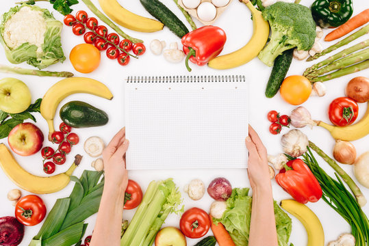 cropped image of girl holding notebook between vegetables and fruits isolated on white