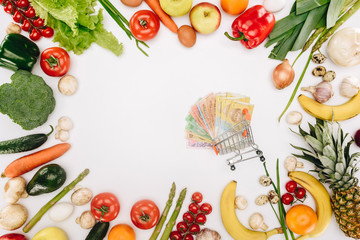 top view of shopping cart with cash between vegetables and fruits isolated on white