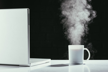 laptop with steaming cup of tea on table isolated on black