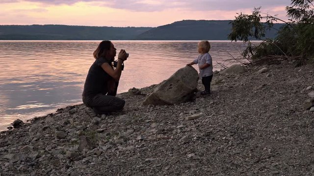 Mother photographing her little child at sunset. Woman with retro camera taking photos of her little son on shore of lake at sunset. Concept of happy family relationship
