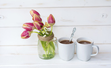 two cups of coffee with milk and bouquet of tulips and lilac on white wooden background