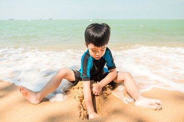A boy in swimming suit is sitting on the beach and playing sand at the sea in the sunny day in summer