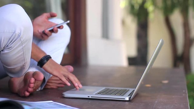 Young handsome man is sitting down on the terrace and using a laptop outside the house.