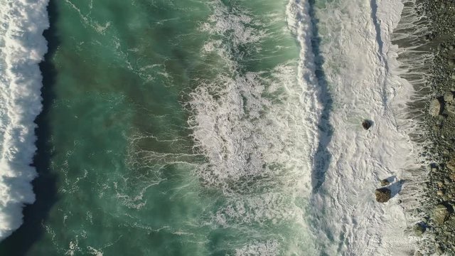 Aerial shot slowly rising while looking straight down at waves crashing on rocky shore. 