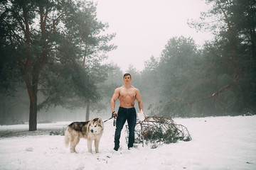 Young bodybuilder with bare torso leads dog Malamute on walk at winter foggy forest.