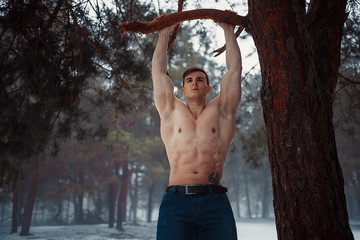 Young bodybuilder with bare torso pulls up on branch in forest in winter. Body hardening.