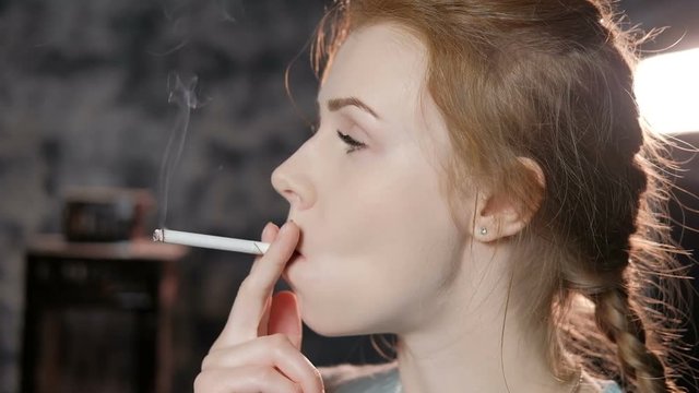 Young woman with red hair in a blue sweater smokes a cigarette in the studio. Slow motion
