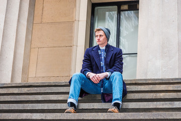 American teenage college student wearing blue long woolen overcoat, scarf, jeans, leather shoes, knit hat, siting on stairs outside office on campus in New York in winter, taking work break..