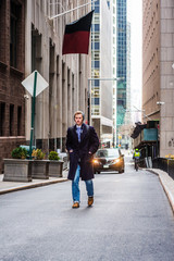 American man traveling in New York, wearing blue long overcoat, scarf, jeans, knit hat, carrying back bag, hands in pockets, walking on vintage street with high buildings. Car, biker on background..