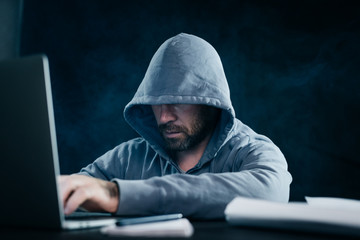 a man hacker in a sweatshirt with a hood sits behind a laptop in a dark room