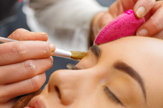 Portrait of beautiful face of young woman getting make-up. The artist is applying eyeshadow on her eyebrow with brush. The lady closed eyes with relaxation, in a blurred background