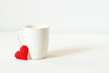 White cup and origami red heart with copy spase. Mockup empty mug on wooden white background. Valentine's day.