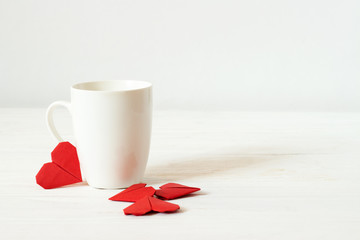White cup and origami red hearts. Mockup empty mug on wooden white background. Valentine's day.